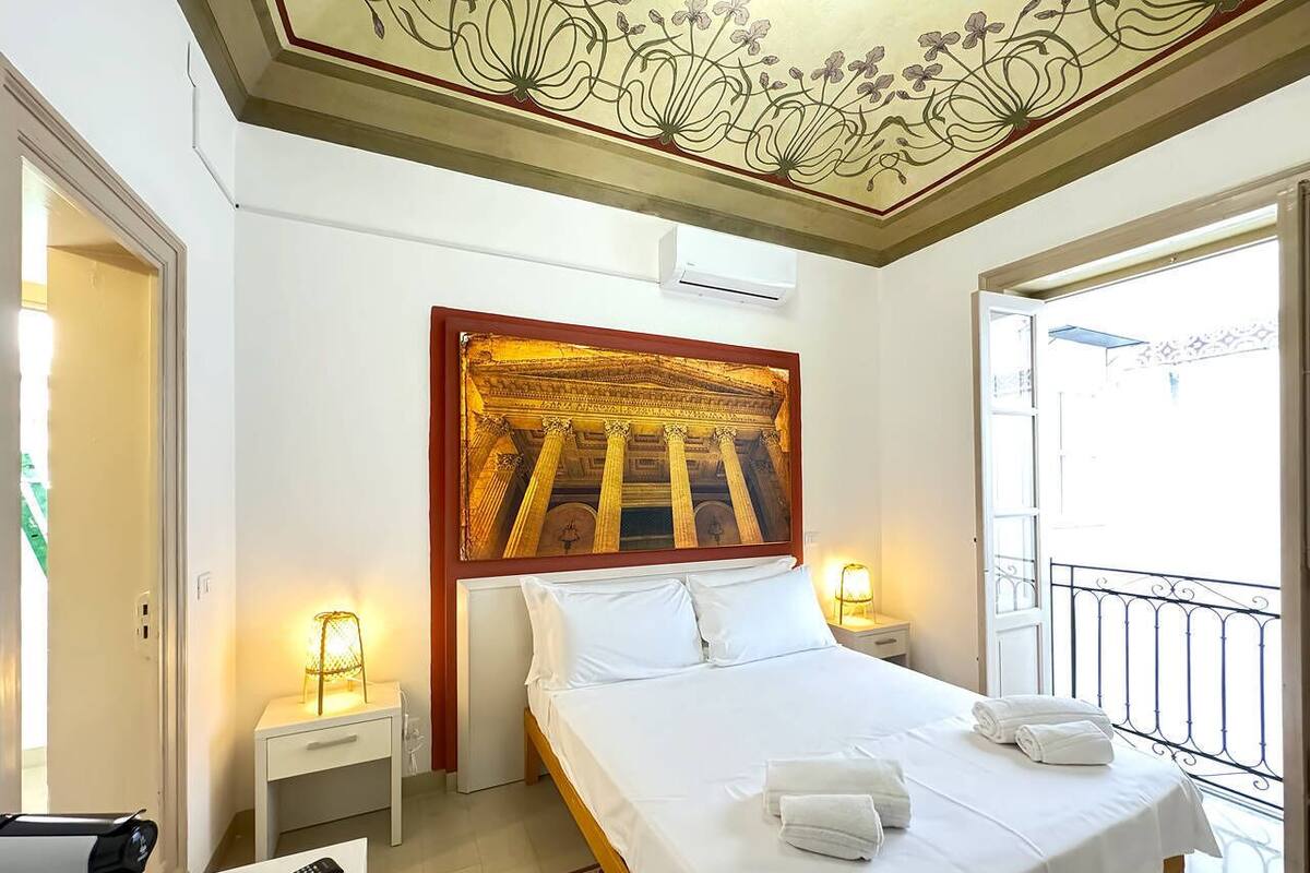 A hotel room with a bed, bedside lamps, a decorative ceiling, and a balcony with a railing.
