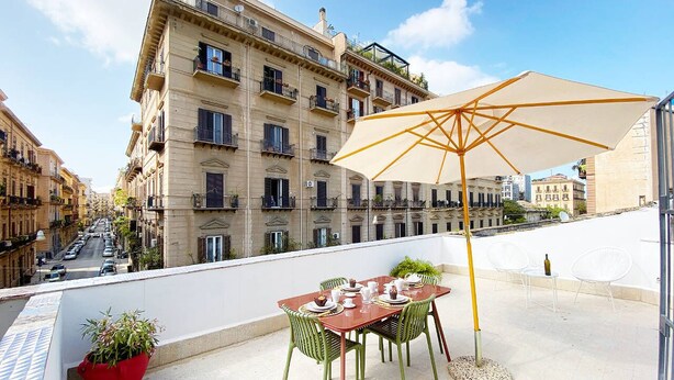 A rooftop terrace with a table set for dining, a large umbrella, and a view of a city street. - Solemar Sicilia - Monacò Rooms (Palermo)