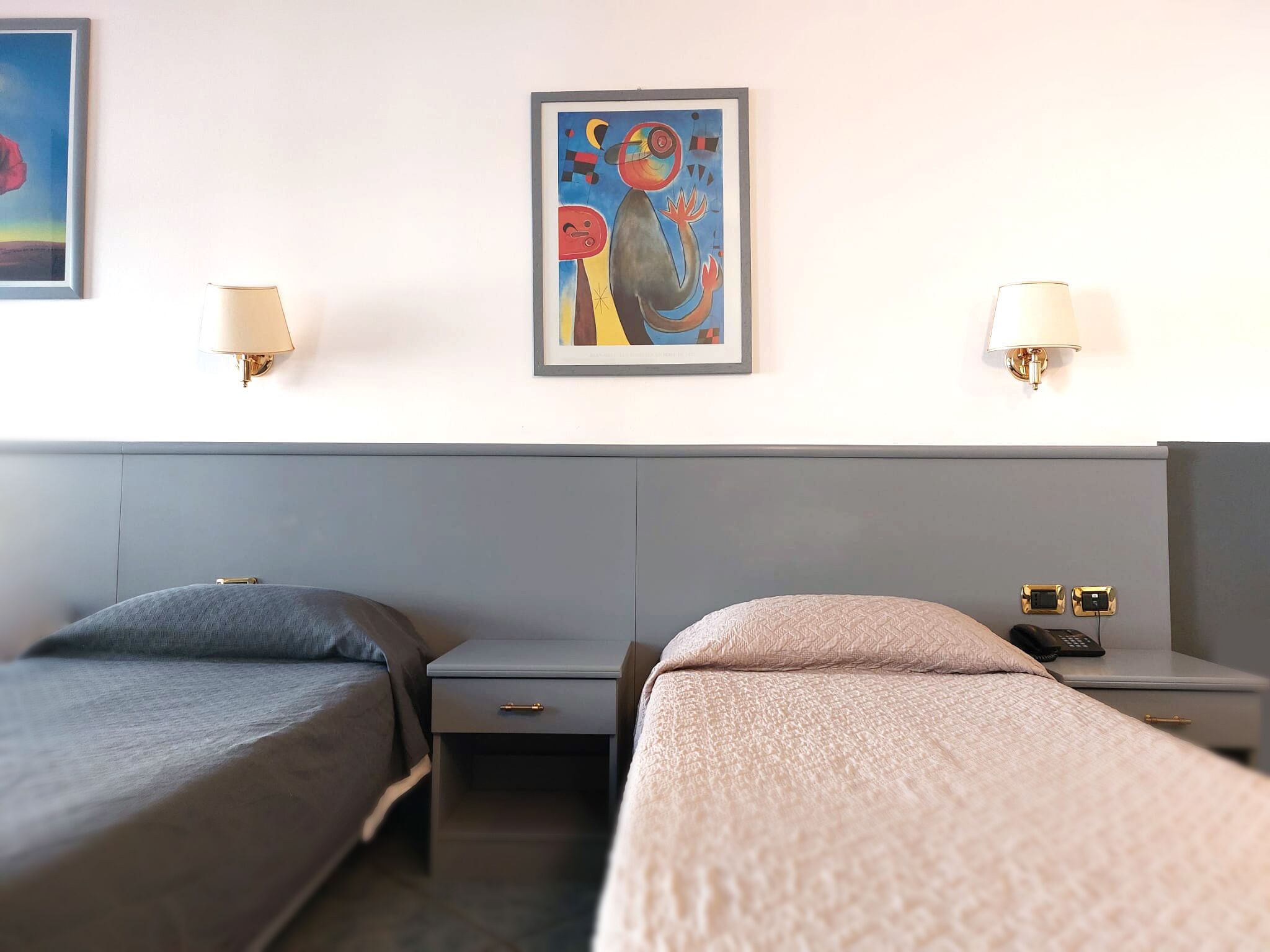 Ein Hotelzimmer mit zwei Einzelbetten, einem kleinen Nachttisch mit Lampe, einem Wandbild und einer Steckdose auf dem Beistelltisch.