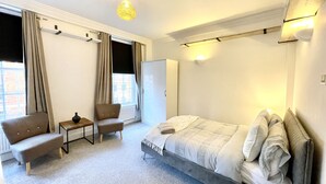 Habitación de hotel moderna con una cama grande, un escritorio pequeño, una silla y un sofá. Cuenta con una ventana con cortinas, un radiador y una lámpara de pared.