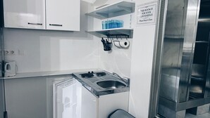 Een ruimtelijk kitchenette met een kookplaat, spoelbak en opbergkastjes. Er staan blauwe glazen op een plank en een bord met een verzoek om netheid.
