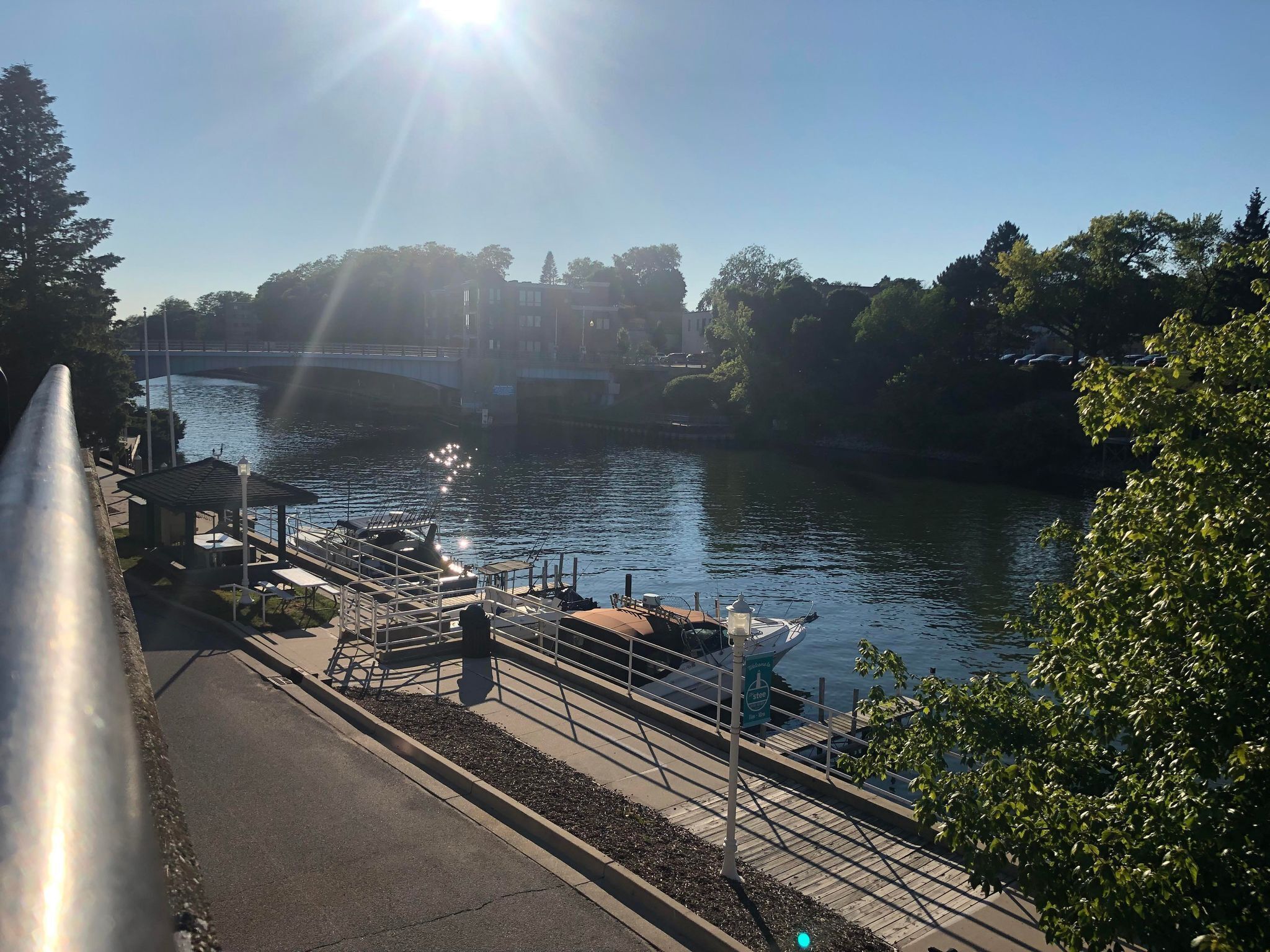 Une promenade le long de la rivière, agrémentée d’un belvédère, d’un quai avec des bateaux et d’un pont en arrière-plan.