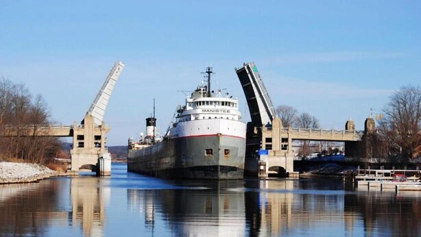 A large cargo ship named MANISTEE passing under a bridge.