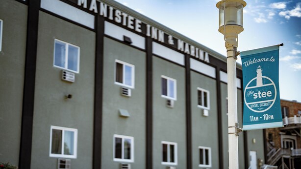 A building with a sign reading "Manistee Inn & Marina" and a banner that says "Welcome to The Stee Social District 11AM - 10PM."