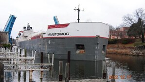 A large cargo ship named Manitowoc docked at a pier with a metal walkway leading to it.