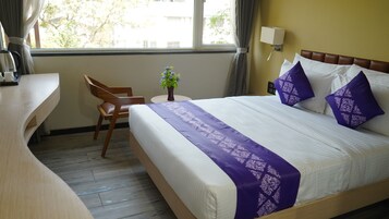 Habitación de hotel con cama, escritorio de madera, silla, mesita con planta y ventana con cortinas.