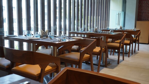 Restaurante com várias mesas e cadeiras, disposto num ambiente moderno com grandes janelas e persianas verticais.