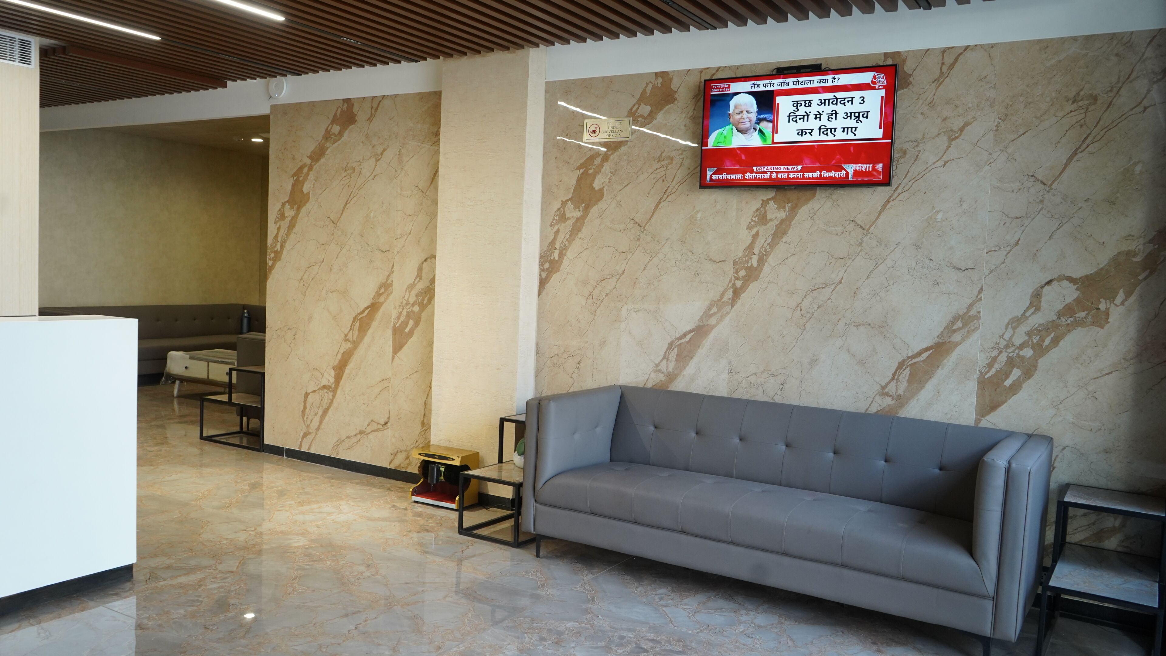 A modern lobby with a grey sofa, marble walls, and a television displaying news.