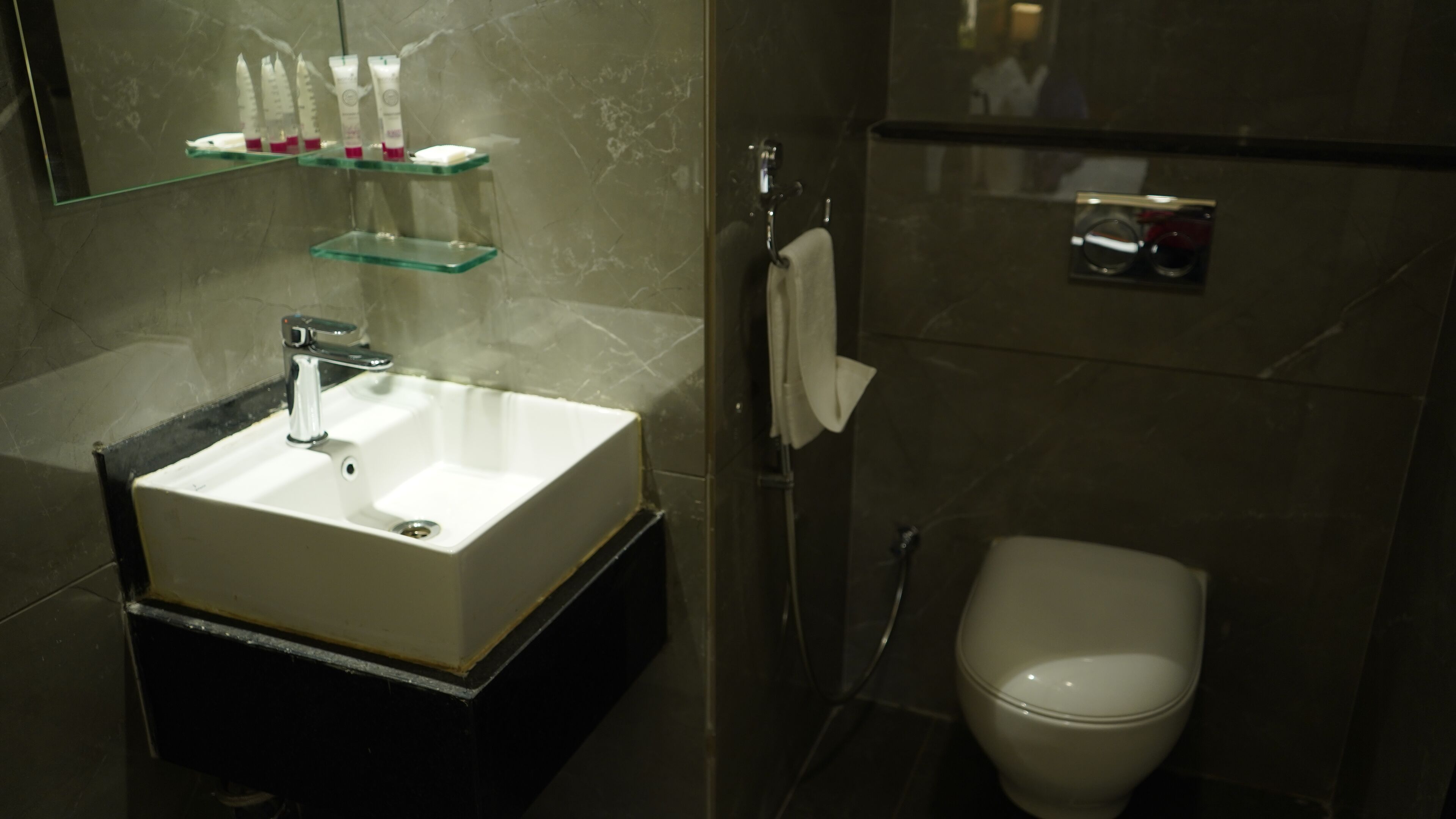 A bathroom with a white sink, a glass shelf above it, a towel hanging on a rack, and a toilet.