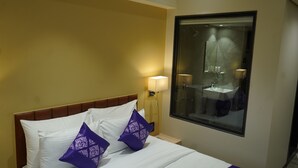 Quarto de hotel com cama, mesa de cabeceira, luminária e janela com vista para um banheiro.