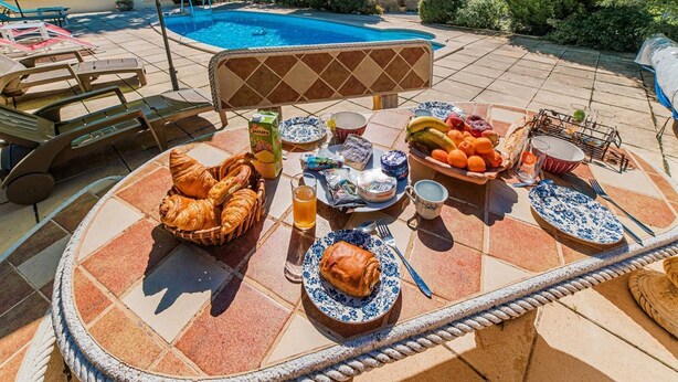 Un cadre agréable au bord de la piscine, avec une table dressée pour un repas, comprenant des croissants, des fruits et des boissons.