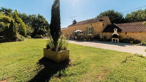 Un bâtiment en pierre avec un toit en tuiles, une allée gravillonnée et une pelouse bien entretenue, avec un grand conifère en premier plan.