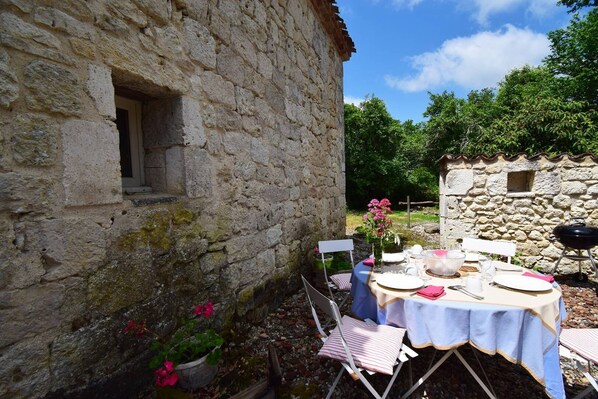 An outdoor dining area with a stone building, a table set for two, and a barbecue grill.