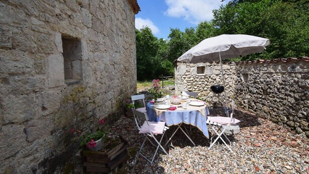 An outdoor dining area with a stone building, a table set for two, and a grill. - Gîte en pleine nature, au cœur du Lot-et-Garonne (LAROQUE TIMBAUT)