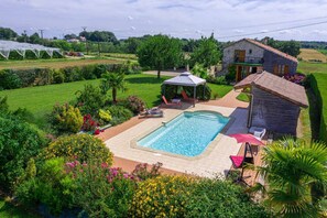 A swimming pool with a patio area, surrounded by lush greenery and a stone house.