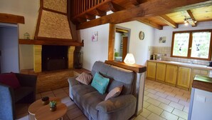 A cozy living room with a fireplace, a sofa, a small table with a plant, and a kitchen area with cabinets and a window. - Gîte avec piscine et jacuzzi - Lot-et-Garonne (BLANQUEFORT SUR BRIOLANCE)
