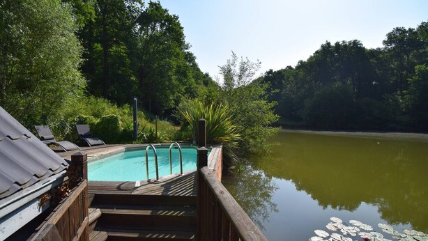 A wooden deck with a pool, surrounded by lush greenery and a calm lake.