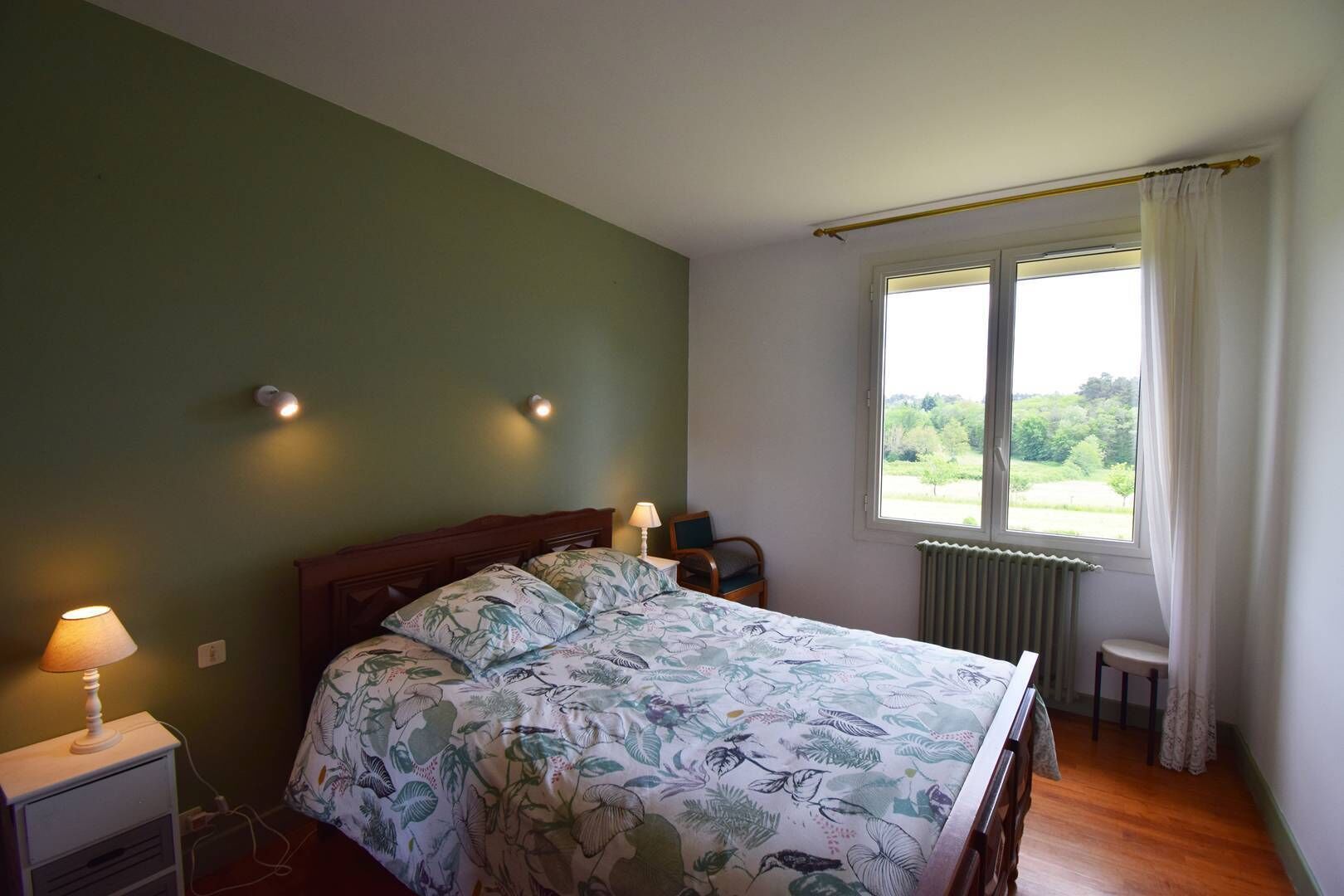 Ein Schlafzimmer mit einem Bett, zwei Nachttischlampen, einem Fenster mit Blick auf Grünflächen und einer Heizung.