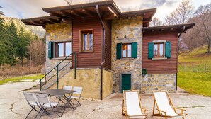 Ein rustikales Steinhaus mit Holzfassade und grünen Fensterläden, eine Beton-Terrasse mit Tisch und Stühlen sowie ein hölzernes Staffelei.