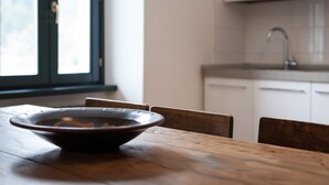Une table de salle à manger en bois, avec un saladier au centre, une fenêtre avec des stores et un coin cuisine équipé de placards et d’un évier.
