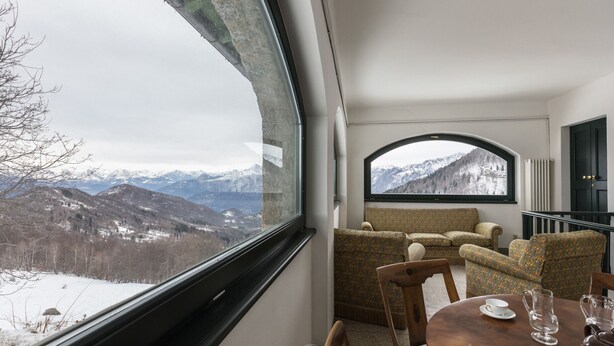 Une pièce dotée d’une grande fenêtre donnant sur des montagnes enneigées. À l’intérieur, on trouve une table en bois avec deux chaises et un canapé à motifs.