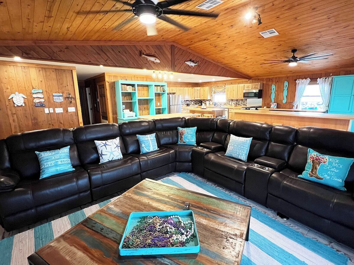 Un accogliente soggiorno con un divano a portafoglio in pelle nera, un tavolino in legno con un vassoio decorativo, e una cucina sullo sfondo con mobili teal e pareti in legno.