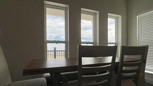A dining area with a table and four chairs, large windows with a view of the sky and a distant structure, and a window with blinds on the right. - White Dolphin - Beachfront - Island Realty (Grand Isle)
