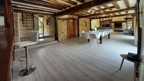 Une pièce spacieuse avec un plafond haut, des poutres apparentes, des murs en pierre, une longue table à manger avec des chaises, un coin bar et une cheminée.