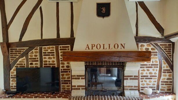 Une cheminée surmontée d’une enseigne portant le nom « APOLLON », située dans une pièce aux poutres apparentes et dont un des murs est en pierre.