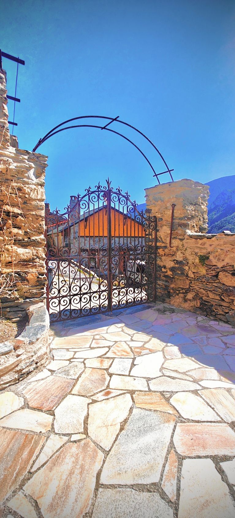 A stone-paved entrance with a wrought iron gate leading to a building with a stone wall and a clear blue sky in the background.