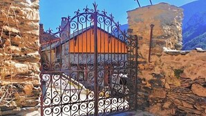 Une entrée pavée de pierres, avec un portail en fer forgé, mène à un bâtiment doté d’un mur de pierre, et un ciel bleu clair se dessine à l’arrière-plan.