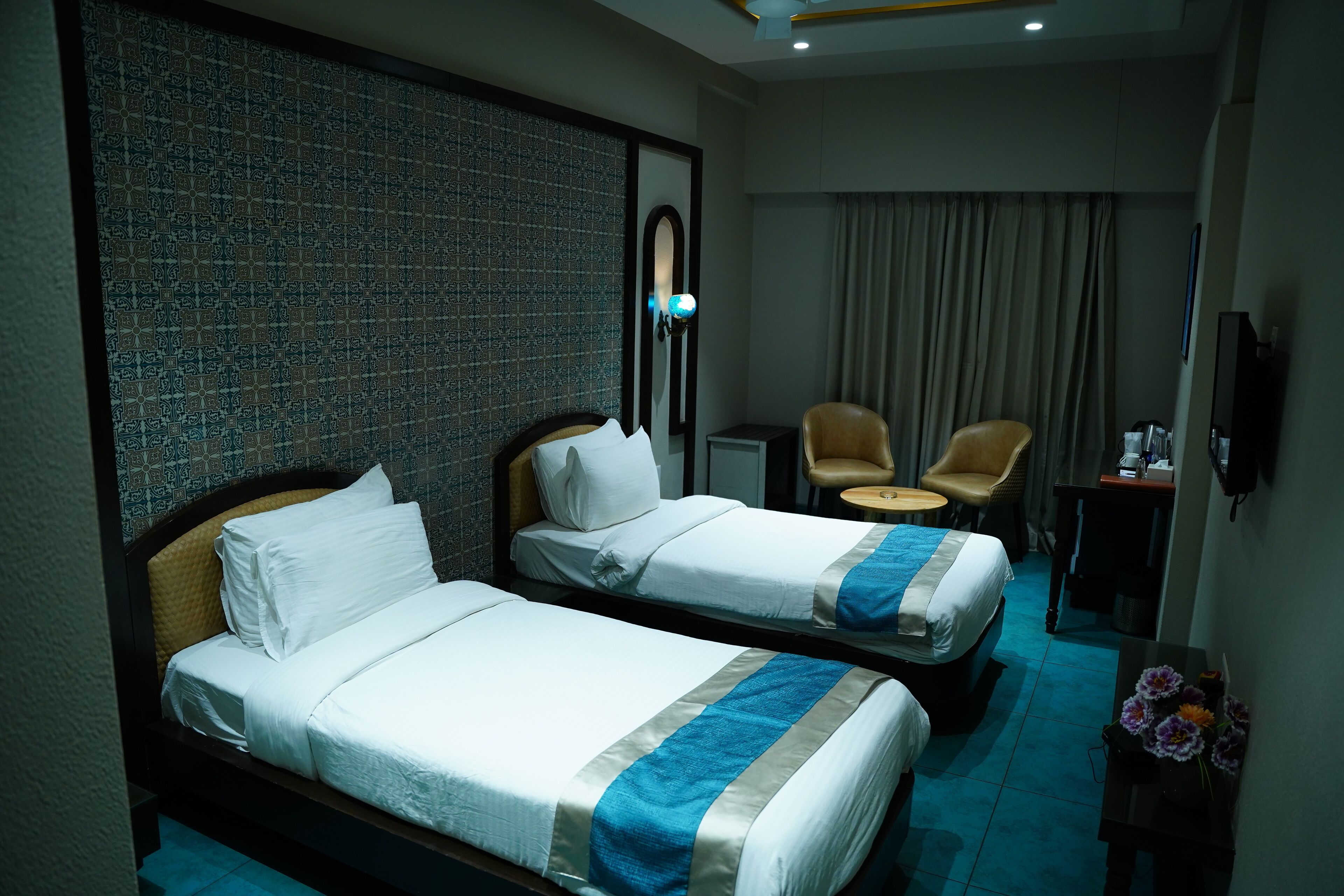 A hotel room with two single beds, a small table, a chair, a desk, and a wall-mounted TV.