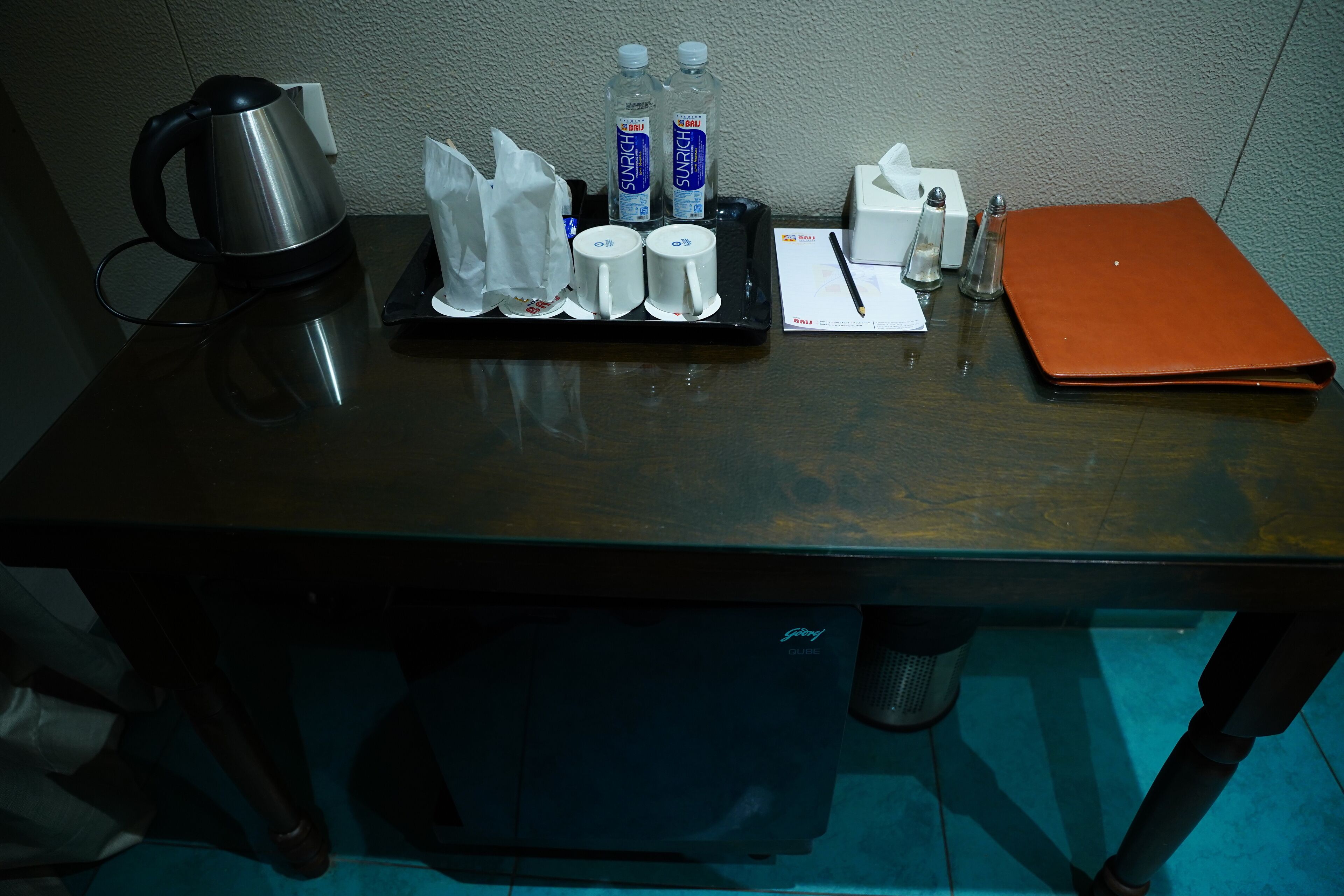 A hotel desk with a kettle, two bottles of water, a tissue box, a notepad, and a pen.