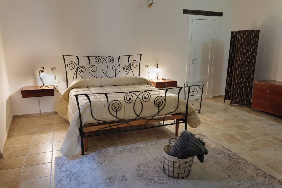 Une chambre à coucher avec un lit en métal, une table de chevet avec une lampe, une commode en bois, une porte et une armoire.