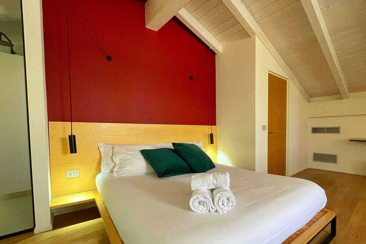 A modern bedroom with a wooden headboard, a red accent wall, and a ceiling with exposed beams.
