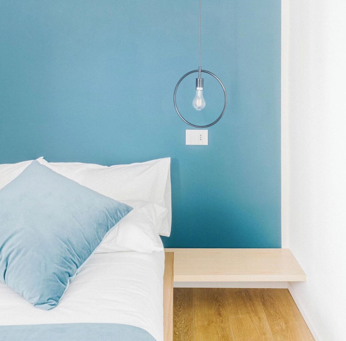 Una cama con ropa de cama blanca y azul, una mesita de noche con superficie de madera y una lámpara fijada a la pared.