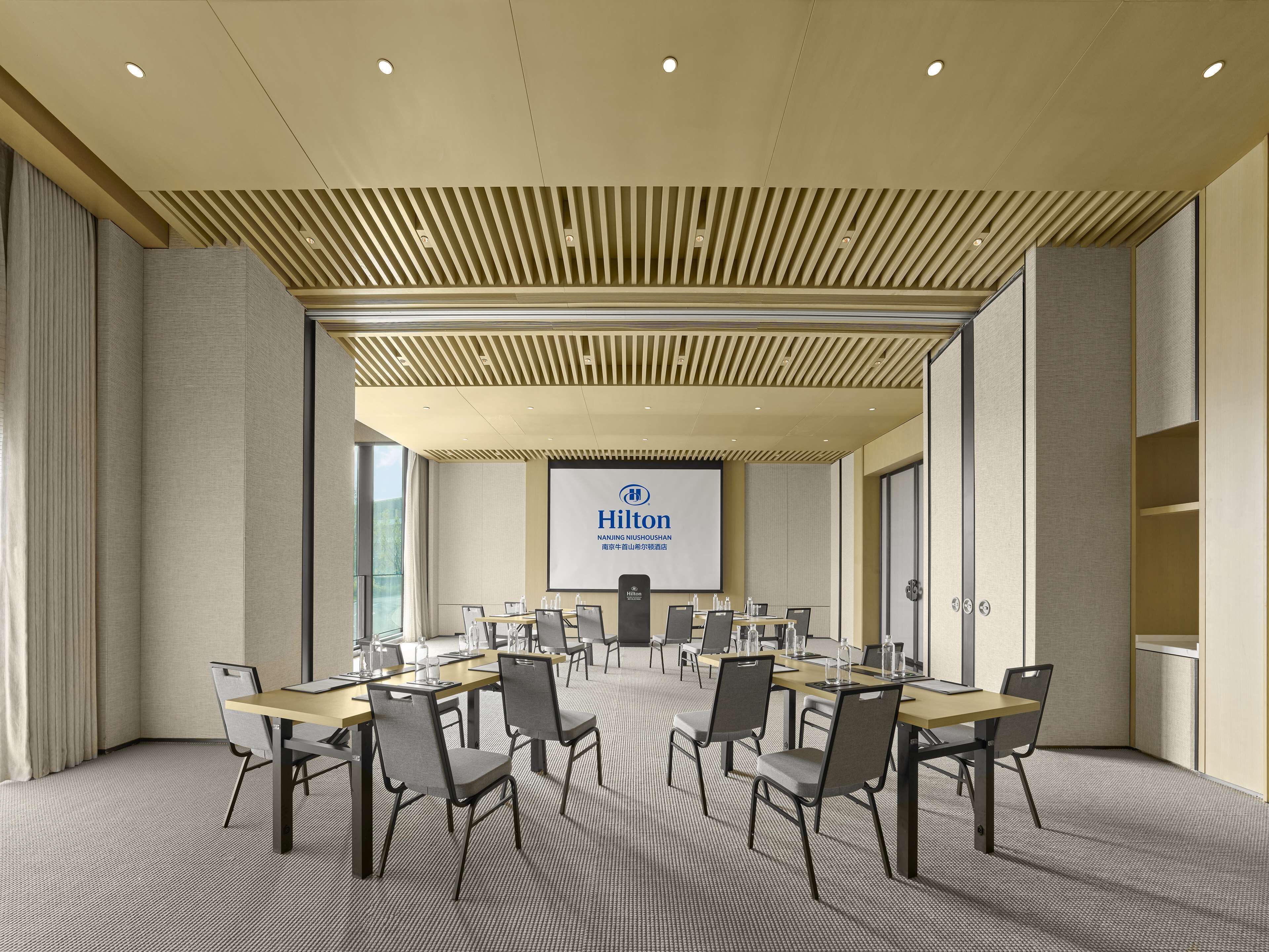 A conference room with a large screen displaying the Hilton logo, multiple tables with chairs, and a modern ceiling design.