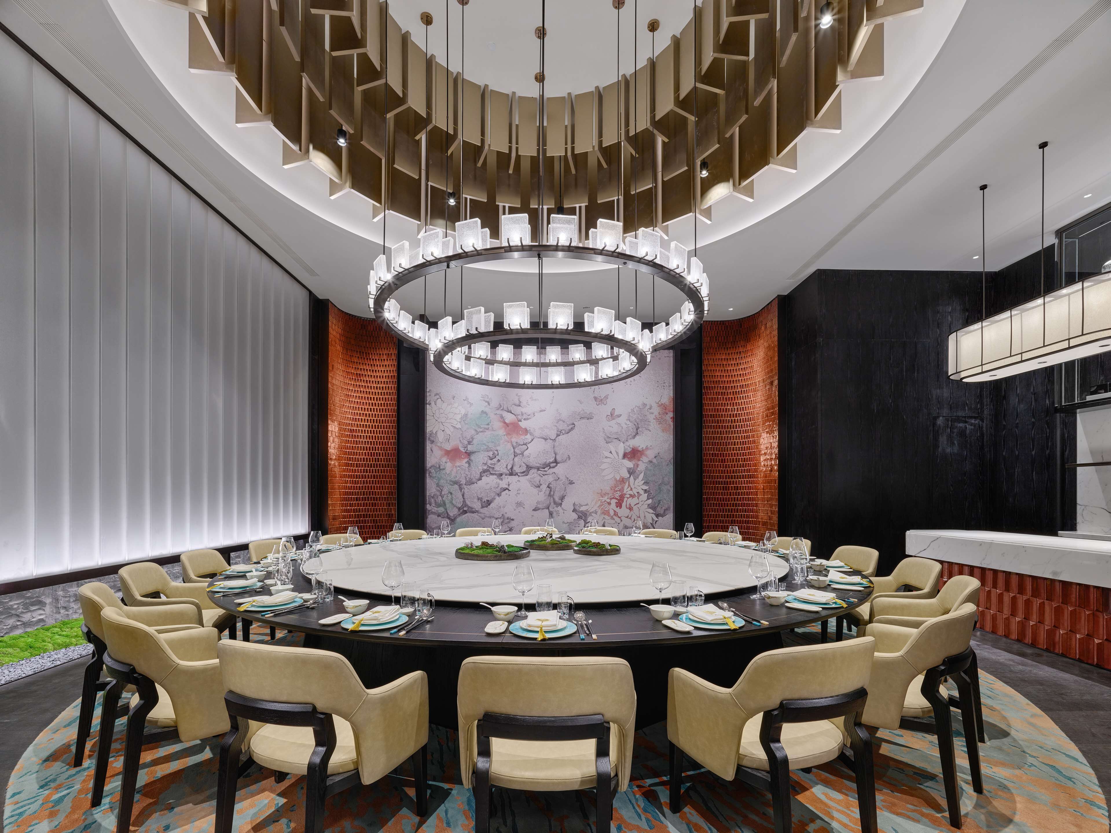 A conference room with a round table, beige chairs, a chandelier, and a marble wall with a floral design.