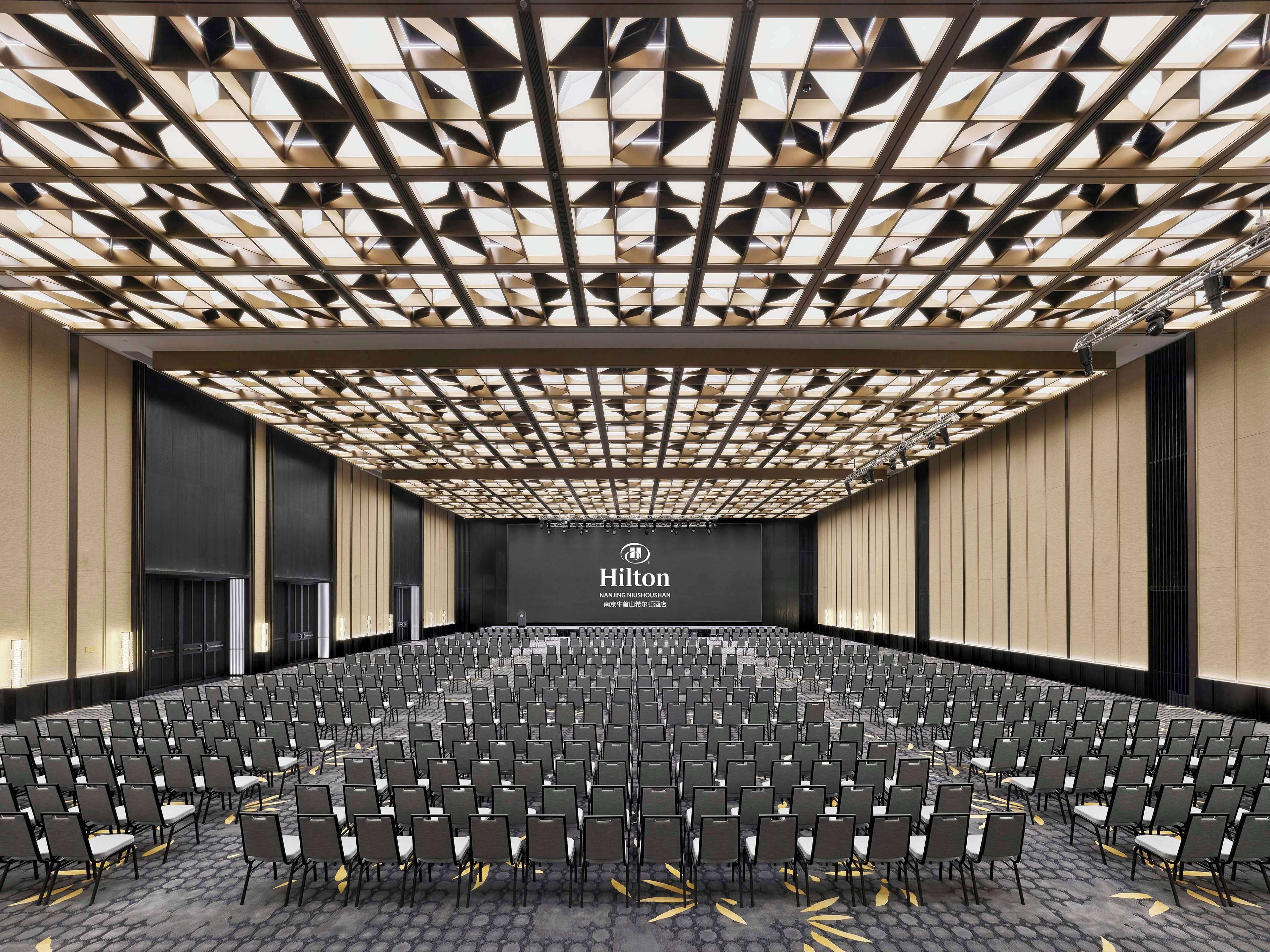 A large, modern conference room with a geometric ceiling design, rows of chairs, and a prominent Hilton logo on the back wall.