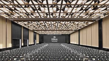 Un amplio salón de conferencias moderno con un diseño geométrico en el techo, filas de sillas y un destacado logotipo de Hilton en la pared de fondo.
