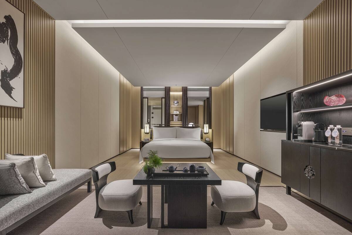 A modern hotel room with a large bed, a dining area with a table and chairs, a TV, and a minibar.