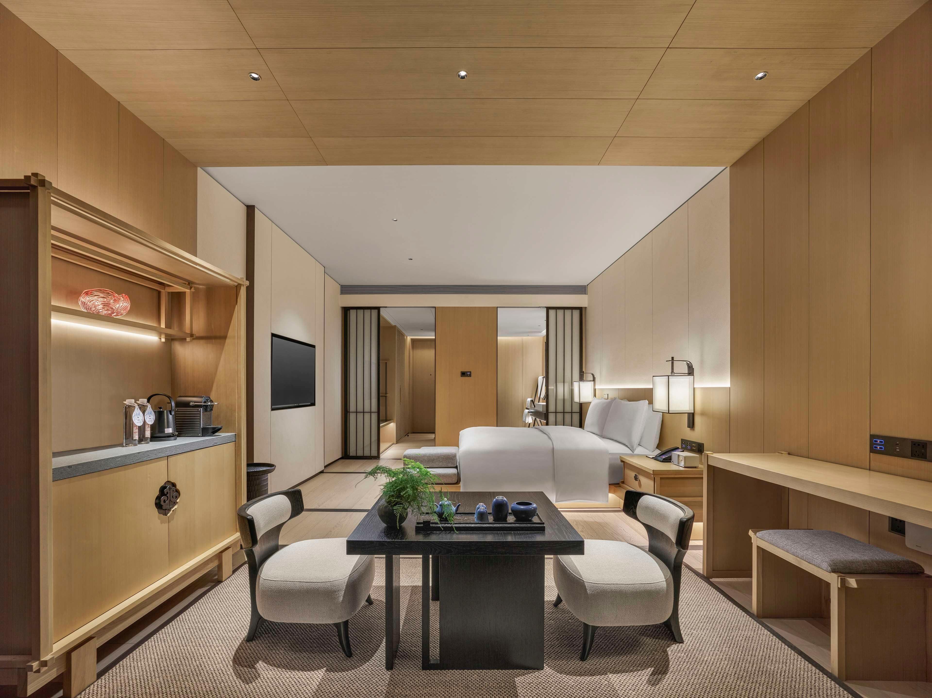 A hotel room with a bed, a desk, chairs, a TV, and a minibar.