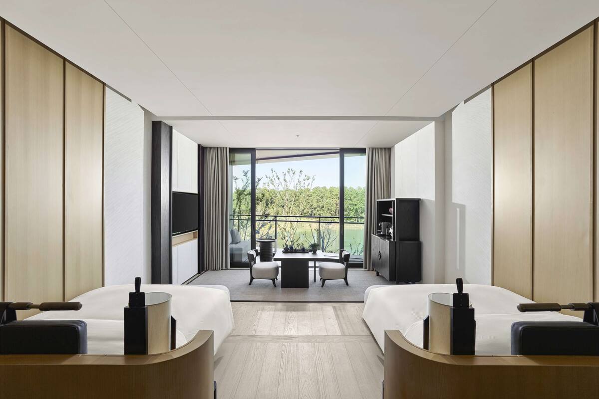 A modern hotel room with two beds, a TV, a desk, and a balcony with a view of greenery.