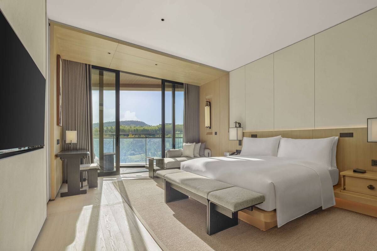 A modern hotel room with a large bed, a bench, a desk, and a view of a forested area.