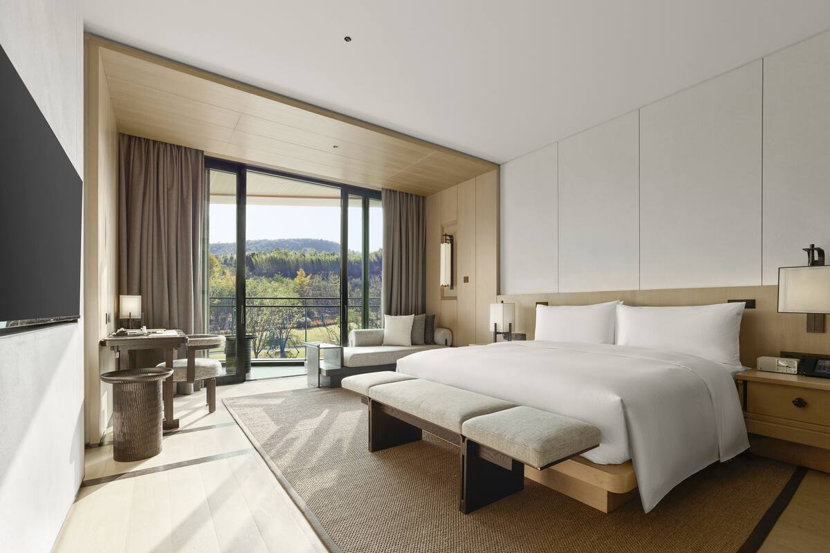 A modern hotel room with a large bed, a desk, a chair, and a view of a forested area.