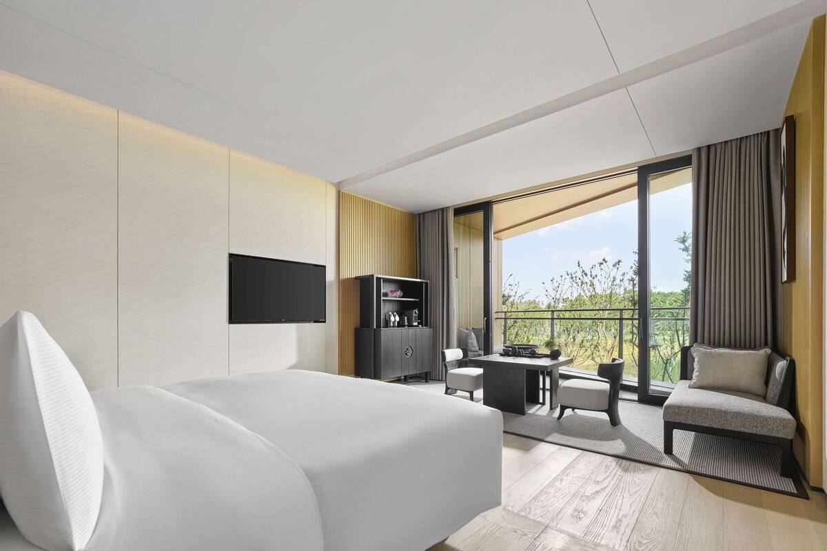 A modern hotel room with a large bed, a desk with a chair, a television, and a balcony with a view of greenery.
