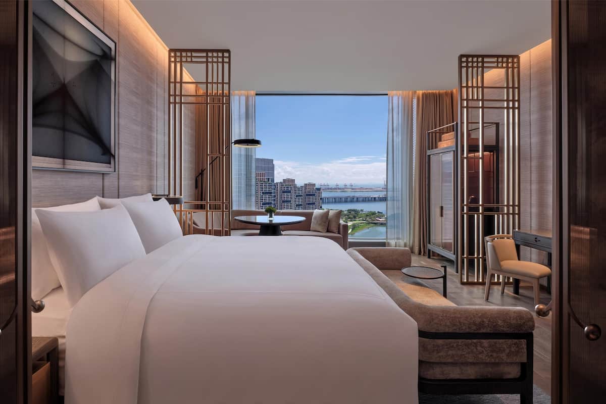 A modern hotel room with a large bed, a round table, a sofa, and a view of a cityscape and bridge.
