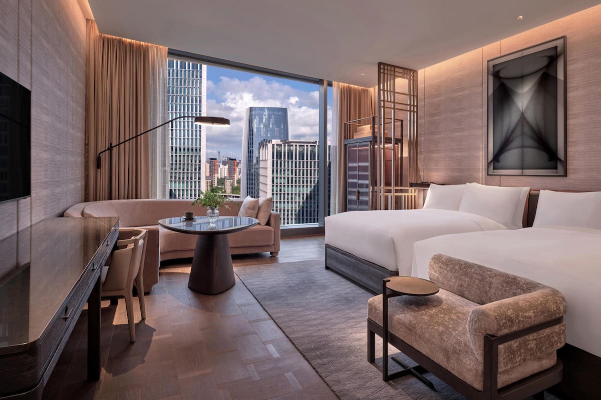 A modern hotel room with a large bed, a sofa, a dining table, and a TV. The room has a large window with a city view.