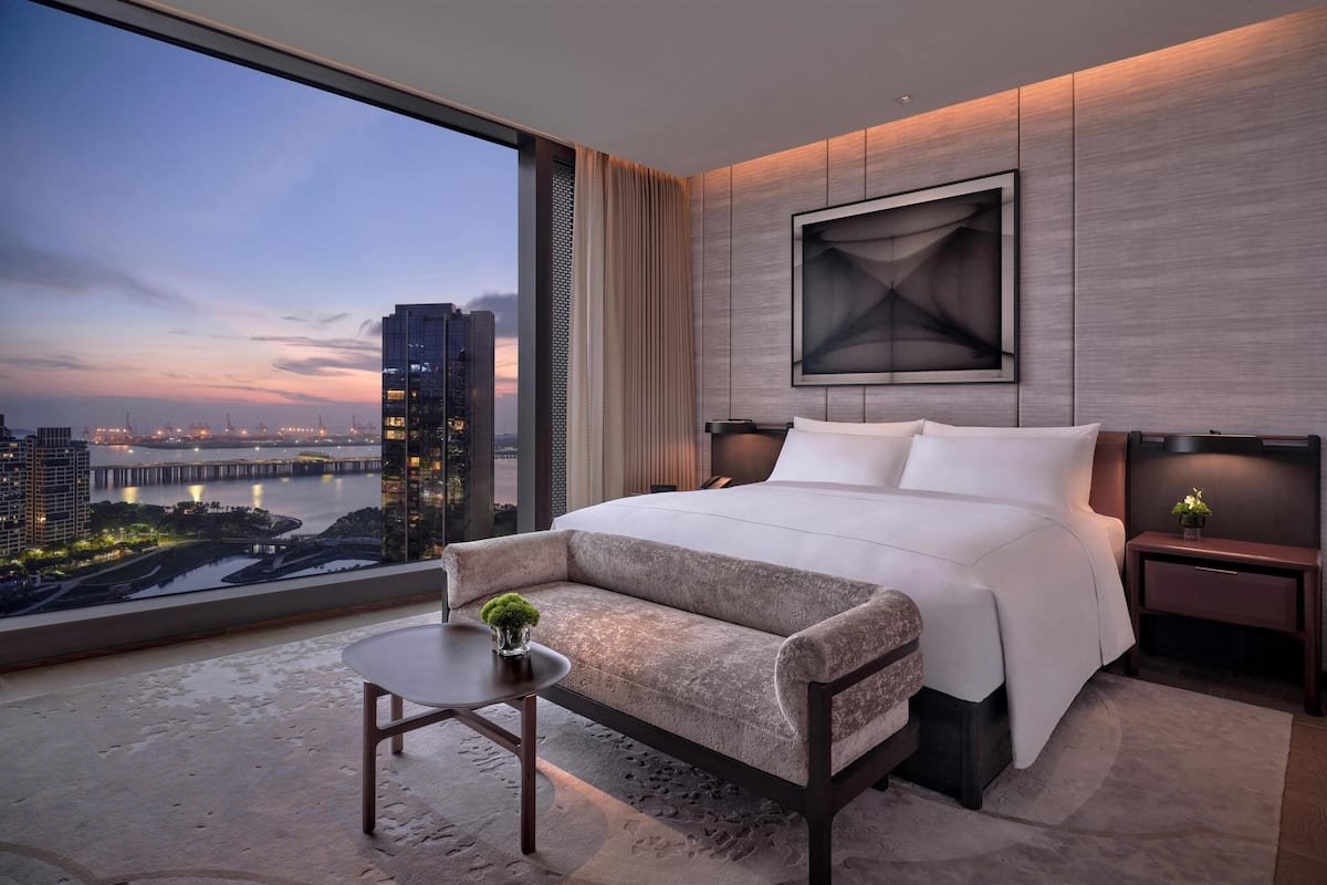 A modern hotel room with a large bed, a sofa, a small table with a plant, and a view of a cityscape and water at dusk.