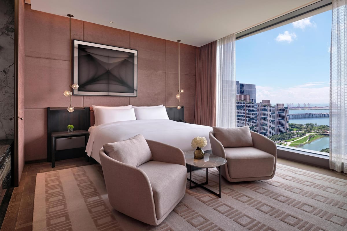 A modern hotel room with a large bed, two armchairs, a small table, and a city view.